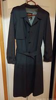 Full view of the men's trench coat hanging on a hanger showing buttons, collar, belt and epaulets.