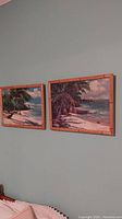 Two framed oil paintings hanging on a wall, showing Hawaiian seashore scenes with palm trees and ocean under cloudy sky.