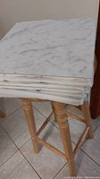 Stack of 42 square peel and stick tiles with white and gray marble pattern, arranged on a wooden stool.
