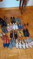 Photo showing an assortment of 18 pairs of various women's shoes arranged on hardwood floor, including boots, sneakers, flats, sandals in multiple colors and styles.