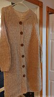 Photo of a hand-knit, long chunky sweater coat with large wooden buttons, hanging on a hanger against a wall and door background.