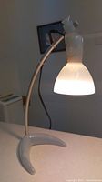 Modern curved desk lamp with white frosted glass shade and base, viewed lit on a flat surface indoors.