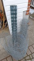 Two wire fencing rolls standing upright outdoors by a wall, one green-coated with square grid, one galvanized chicken wire.