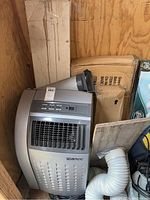 Portable TechniTrend air conditioner positioned on wooden floor inside shed, with flexible vent hose attached and cardboard boxes behind it.