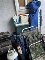 Side view of lot showing stacked coolers, propane grill, propane tank, folding chairs with camouflage fabric, blue canopy bag, and outdoor setting.