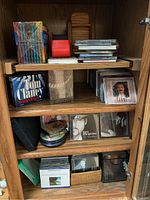 Wide view of a wooden cabinet shelf containing an assortment of CDs, audiobooks, and CD storage cases. Visible are music CDs including artists Toni Braxton and Tim McGraw, John Grisham audiobook, and a 10-pack of Maxell CD jewel cases.