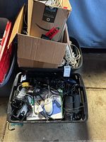 Black bin packed with various cables, electronic devices, and boxes. Visible Amazon Prime cardboard box, cables tangled inside.