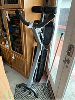 Folded exercise bench with padded backrest and seat cushions leaning against a wall near a wooden cabinet and glass door.