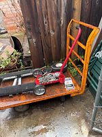 Orange flatbed cart loaded with a red roller dolly holding a metal chain, two tires, and a black metal ramp or platform near a wooden fence.