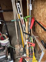 Garden tools including shovel, pick, hand rakes, edger, and string trimmer stored in a shed against a wooden wall.