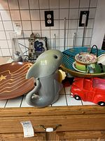 Full view of ceramic dolphin paper towel holder, red toy truck, blue bowl, lighthouse cookie jar, and other pottery items on a tiled counter.