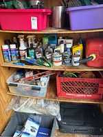 Wooden shelves containing various outdoor hardware, lubricants, cleaning supplies, oil bottles, hand tools, electrical cords in a red crate, gas container, and assorted hardware in plastic tubs.