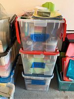 Three stacked large plastic bins filled with office supplies, some with red lids and blue labels marked 'Office'.
