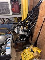 Paint sprayer unit with hoses stored in wooden shed alongside yellow metal horses and other stored items.