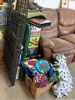 Wide shot showing folding metal dog cage leaning on couch, boxed folding pet ramp behind it, and large box containing various dog toys and supplies.
