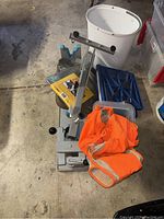 Shooter vice with orange safety vest, white bucket, blue foldable stool, and other assorted garage items piled together on the floor.