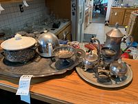 Overview of all silverplate items displayed on kitchen counter, including large serving platter, covered serving bowl, footed bowl, and tea service on round tray