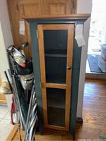 Front view of small wooden display cabinet with glass door, showing two shelves inside and natural wood frame around glass.