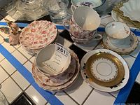 Multiple Spode Rosebud china pieces including plates, cups and saucers arranged on a surface with blue tape edges. Includes floral patterned plates and gold-trimmed saucers.