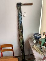 Full view of the tall wood saw standing vertically, showing the overall painted blade and wooden handle with metal fitting.