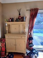 Front view of the tall painted wood cabinet with objects on top, showing upper door and lower drawer plus double doors on base, positioned near a window with curtain.