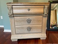 Front view of two-drawer wooden cabinet with decorative metal handles, light wood finish, and ornamental corner accents.