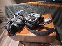 Three cameras on wood table: Kodak EasyShare CX7330 digital camera, Canon digital camera DS126191 in its case, Sony video camera in its case and user manual.