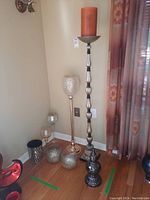 Tall silver and black stand candle holder with orange candle, alongside other metallic candle holders and glass vases in corner.