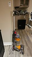 Full view of upright Dyson DC24 vacuum cleaner standing upright beside a wall and furniture.