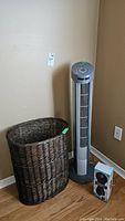 Woven laundry hamper basket and Seville Classics tower fan side by side against a beige wall on a wood floor.