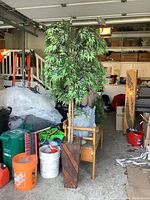 Full view of the faux tree with green leaves standing in the wooden planter among other stored items in a garage.