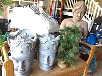 Two ceramic head plant stands and the resin lady statuette on a wooden surface with a small fake fir tree.