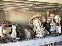 Shelf display of garden art including ceramic planter, resin frog welcome sign, terra cotta girl head, wood figurine, and lanterns