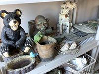 Display of black wood-look bear figurine sitting with a blue bird on a planter bowl, boot-shaped planter with green frog, white resin dog figurine, and several flat decorative wall plaques on the shelf.