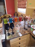 Overview of multicolored goblets, black glasses, cocktail glass, clear stem glasses and lamp on countertop