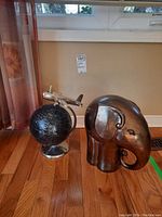 Two decorative items against a beige wall and window: large wooden elephant figurine and black globe with airplane.