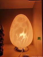 Egg-shaped glass lamp illuminated, showing its opaque white mottled glass and metal base from an angled front view.