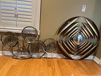 Wider shot showing the two metal wall art pieces positioned on the floor against a wall with hardwood flooring; one piece with five metal wheels arranged horizontally, the other a large circular piece with layered metal strips and a mirrored center.