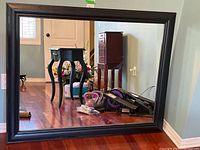 Front view of the large rectangular black framed mirror showing clear reflective surface and wood floor reflection.