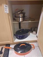 Photo of cabinet shelf with stainless steel pots, lids, blue enamel pot, and orange pan on floor.