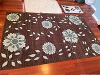Photo of large floral patterned brown area rug with light gray and white flowers and greenish leaves on wooden floor, part of two smaller mats in view.