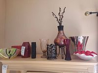 Full view of collection showing various glass vases and bowls including a green-black bowl, red vase, textured black vase, beige-gold patterned vase, metal candle holder, large multicolored vase on stand, tall purple vase, tall reflective metallic vase and ruffled white-red bowl.
