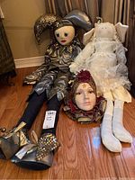 Two large dolls sitting on a hardwood floor, one dressed in metallic silver jester outfit with a ceramic head, the other in white lace angel dress.