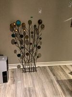 Metal wall art standing against a gray wall on light wood flooring, showing colorful disks and mirror parts on slender metal branches.