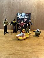 Photo showing five Mcfarlane Beatles figures, miniature Yellow Submarine, clown figure, and Beatles collector's book.