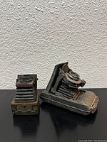 Front and side view of two antique camera bookends shaped like vintage folding cameras, showing intricate details and worn finish.