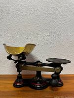 Side view of antique cast iron balance scale with large brass weighing pan and small iron weighing plate showing black body with red painted details.