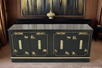 Front view of a black vintage Asian style credenza with gold painted scenic designs and brass handles, placed in a room with a wooden floor and dark background.