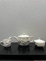 Three-piece ivory tea set including a teapot, creamer, and covered sugar bowl with raised floral motifs on black surface against textured wall.
