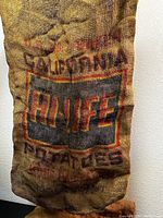 Close-up of a vintage burlap potato sack printed with 'CALIFORNIA HI-LIFE POTATOES' in black and red ink on brown jute fabric.